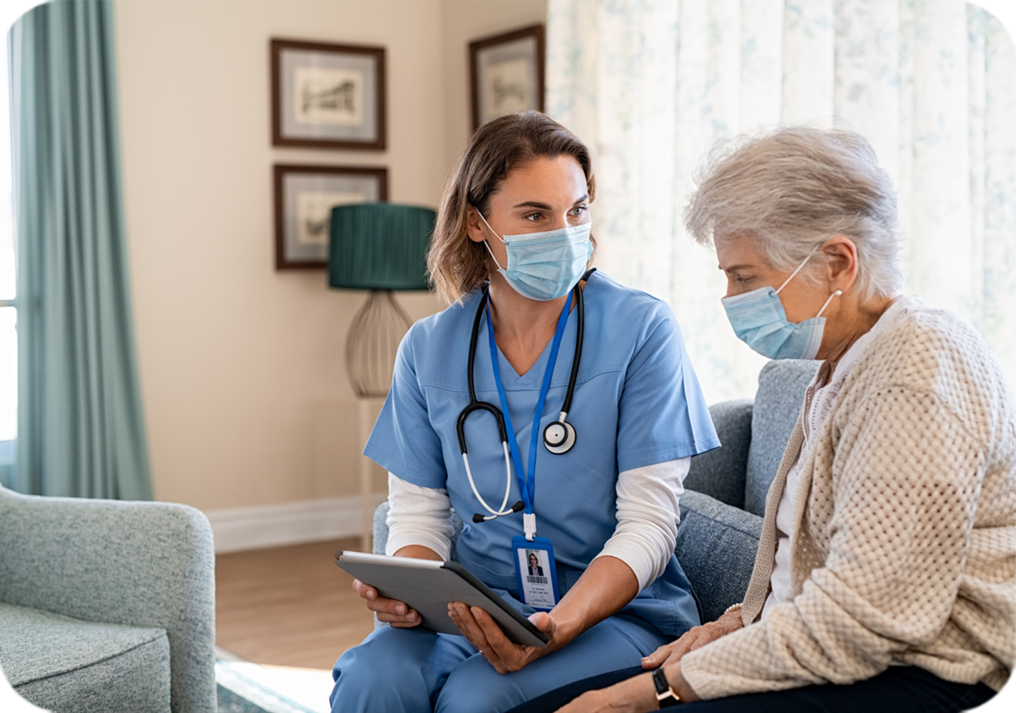 medical professional in scrubs seated on couch next to older woman, both looking at tablet